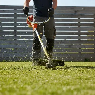 A Husqvarna 128LD gas string trimmer in action, trimming grass along a fence line with clear debris path.